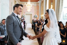At Danesfield House, a wedding photographer captures a poignant moment. The image, shot from behind the altar, shows the couple holding hands and smiling during their ceremony as their seated guests watch on.